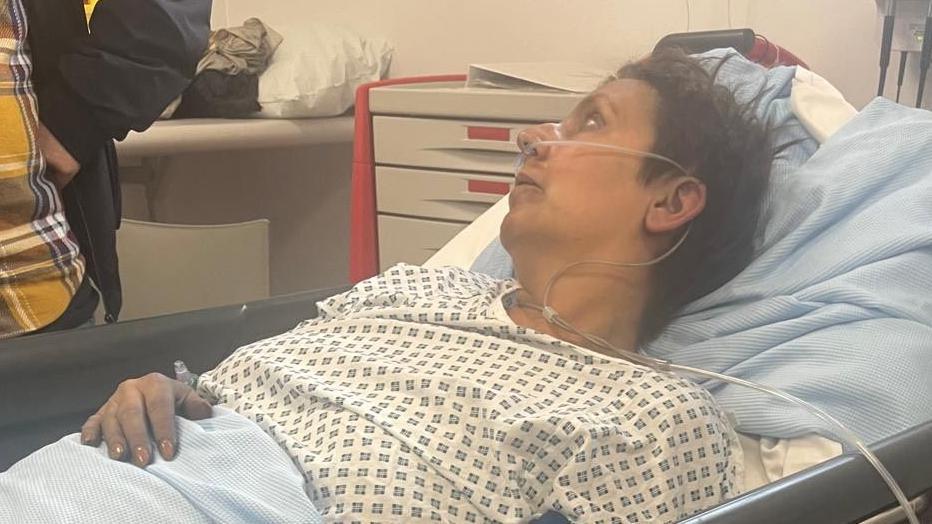 Emma Martin lies in a hospital bed in a hospital with a tube attached to her nose. She is suffering has smoke inhalation from a fire and has two broken ankles and a broken vertebra in her back after jumping out of a first-floor window. She is captured from the side and talking to a man wearing a yellow and black checked shirt whose head and shoulders are out of shot.