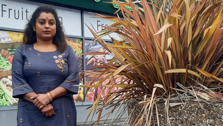 Thilakshana Shanthakumar standing outside her shop beside a large planter with a spiky brown cordyline plant in it