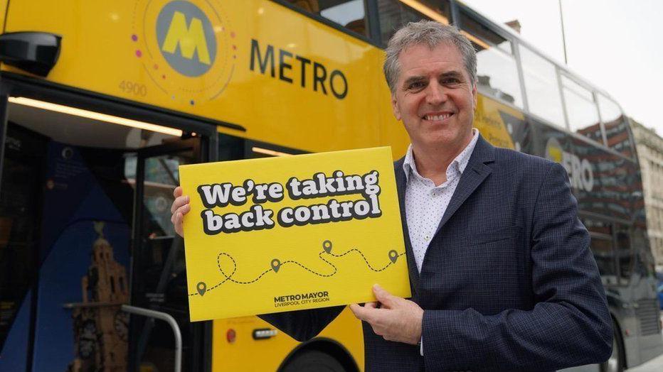 Steve Rotheram, with grey hair and wearing a navy blue blazer, smiles at the camera in front of a yellow bus holding a sign saying 'We're taking back control'. 