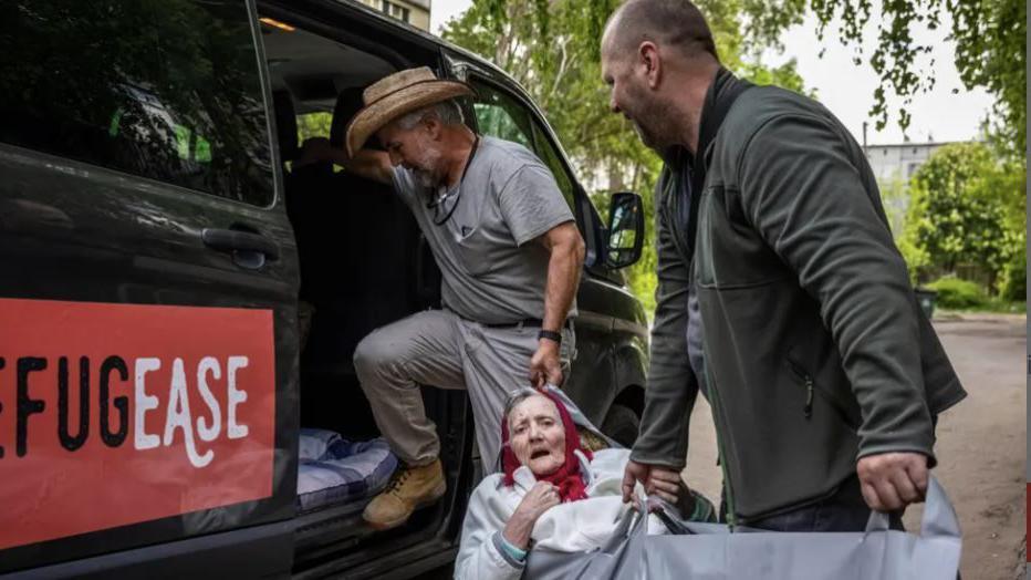 Two volunteers for Refugease carry an elderly woman into a black aid vehicle with the red Refugease logo on the side