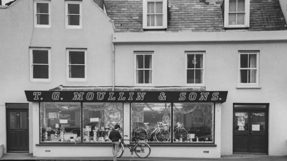 A black and white photograph of the bicycle shop in Guernsey. The signage of the front of the shop has the words 'T.G. Moullin & Sons'. There is a bicycle out the front.