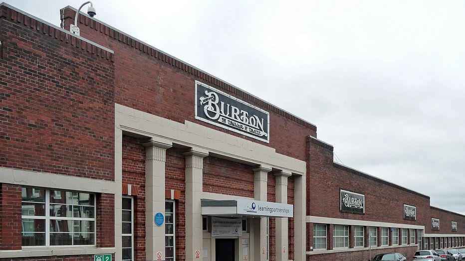 A large redbrick factory with white columns at the front and a tiled sign reading 'Burton the tailor of taste' in black and white above the entrance