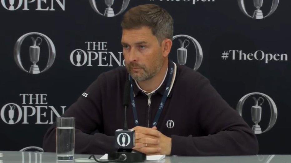 Chief executive of the R&A Mark Darbon is sitting at a microphone and has a glass of water nearby. He is in a black jacket with a blue lanyard on. He is standing in front of black wall with The Open insignia visible