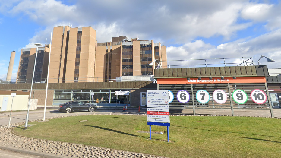 The main entrance to Raigmore Hospital. There is a sign on an area of grass and behind it the numbers and colours of different zones inside the hospital.