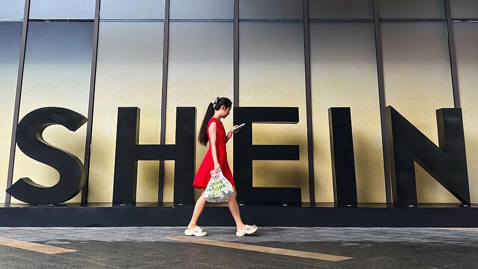 A woman in a red dress looks at her smartphone while walking past a Shein sign outside the company's office in Guangzhou, China. 