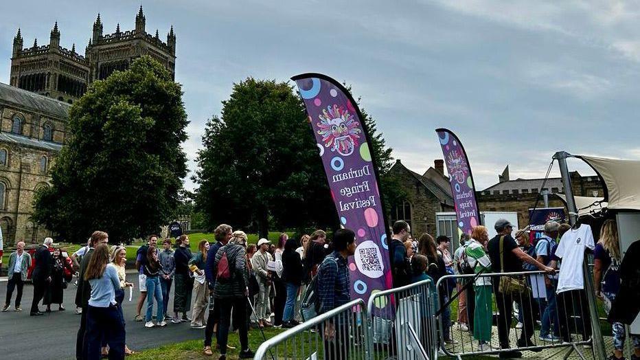 People queueing outside a Durham Fringe tent on the city's Palace Green outside Durham Cathedral in July 2023. there are purple Durham Fringe banners.
