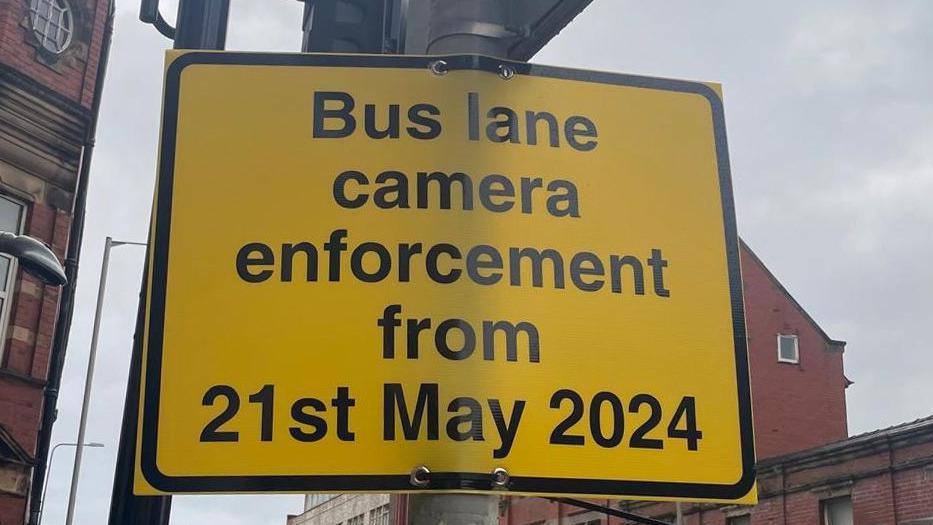 Close up of a yellow street sign with black writing on a lamppost saying 'bus lane camera enforcement from 21st May 2024'. There are red brick buildings in the background and a grey, cloudy sky