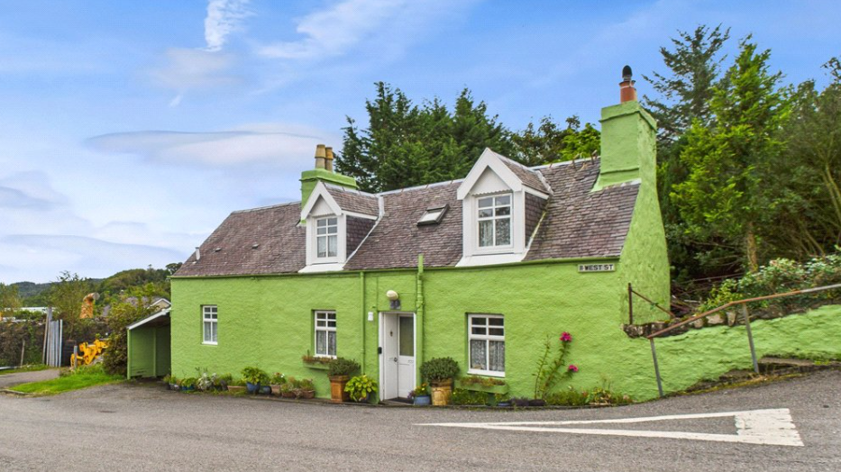 Balamory's green house on West Street, a two-storey cottage in Tobermory, has a slate roof and flower pots outside. the road outside leads downhill to the harbour, where a ferry can be seen. 