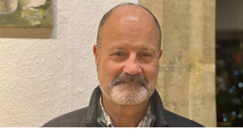 Brian Freeston, a bald man with a grey beard and wearing a checked shirt and brown jumper, looks straight into the camera.