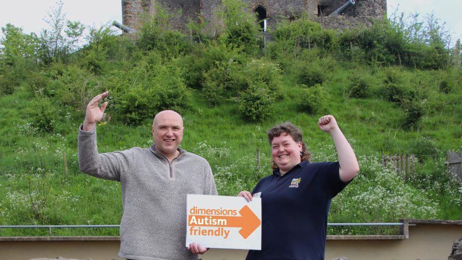 Two people hold an autism friendly sign in front of a castle