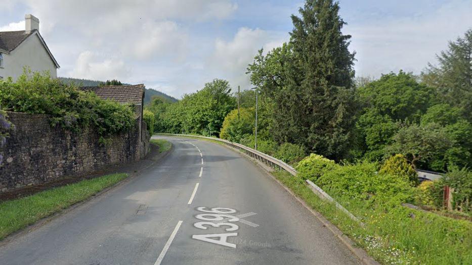 A view of a bend on the A396. The road is lined by trees and a wall. 