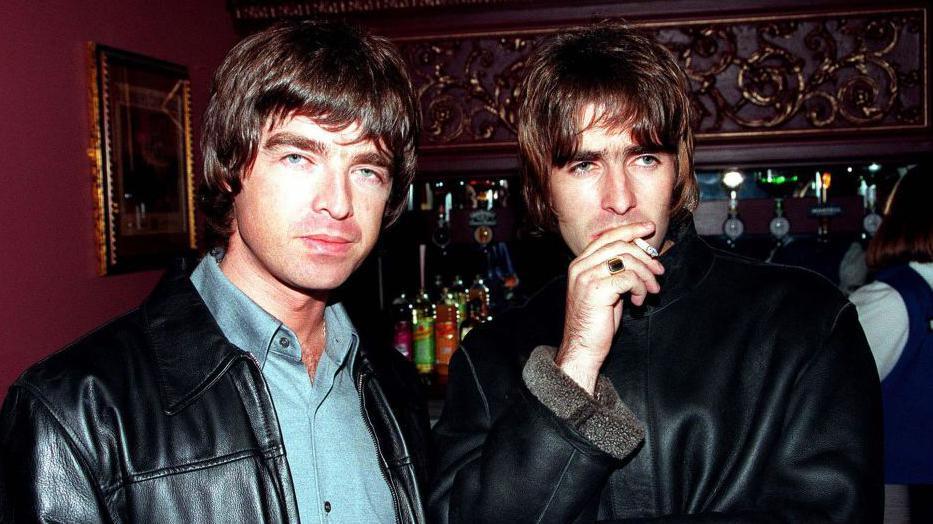 Picture of Liam and Noel at the opening night of Steve Coogan's comedy show in the West End, London in 1995. They are stood together with a bar behind them. Liam, on the left, is wearing a blue shirt and a black leather jacket and is looking at the camera. Noel, on the right, is looking away from the camera and is holding a cigarette up to his mouth. He is wearing a black leather jacket.