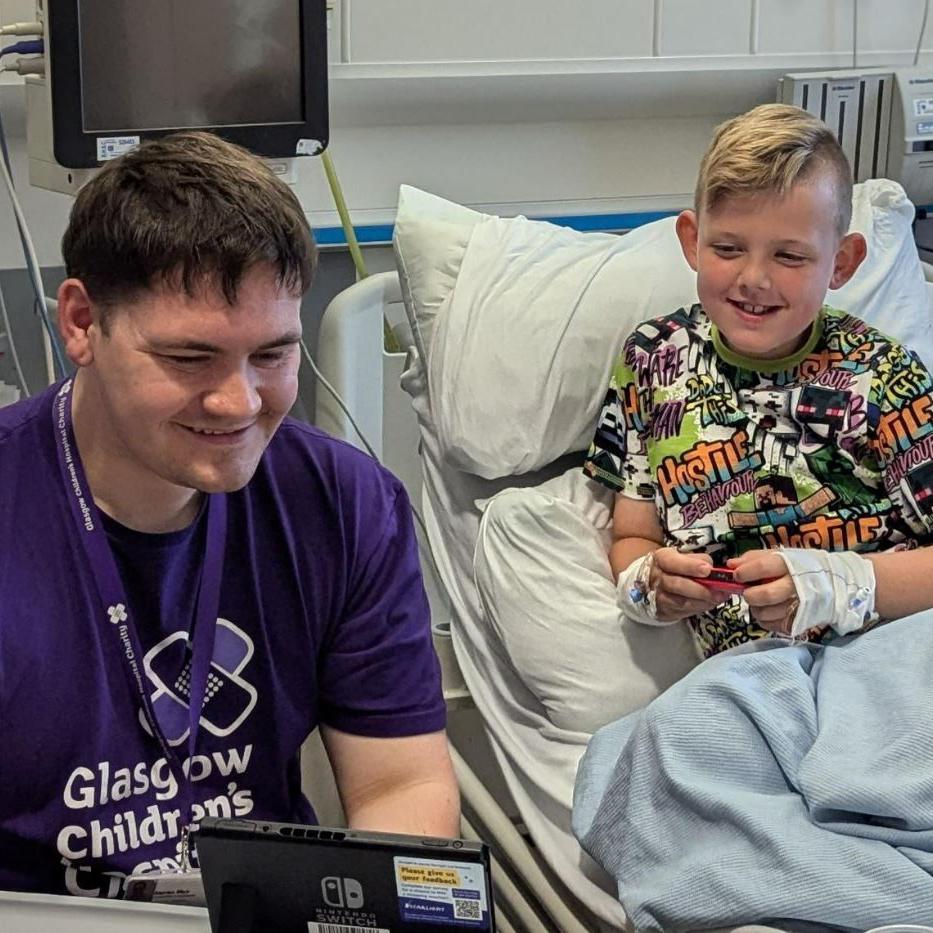 A young boy in a hospital bed games with the Gamer-in-residence. 