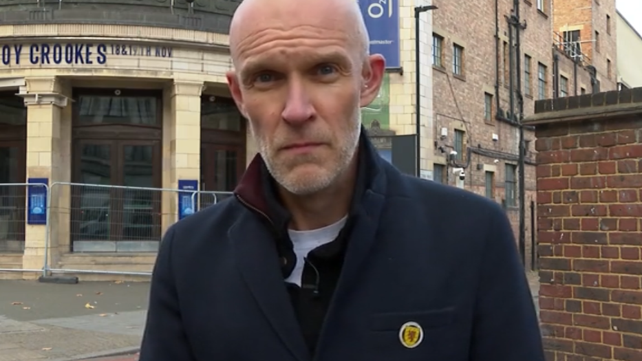 A bald man in a navy coat with a badge with the Scotland crest on it stands in front of the Brixton Academy in London. A sign reads "Joy Crookes" over the entrance.
