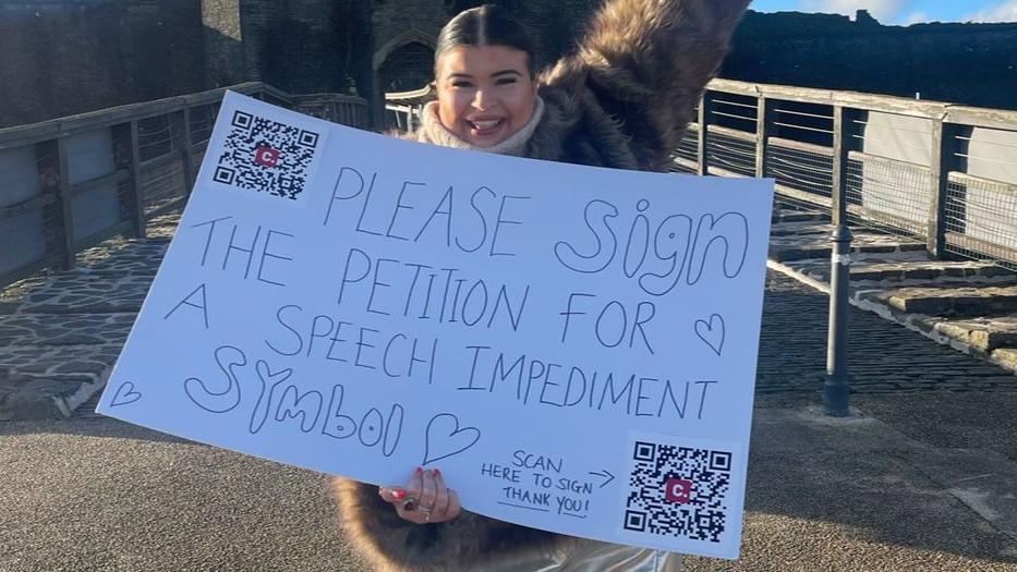 Jessie Yendle, who is grinning and holding her campaign poster which says: Please sign the petition for a speech impediment symbol. 