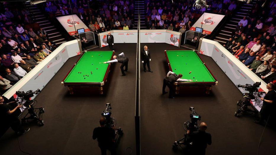 An overhead view of the Crucible Theatre's stage, surrounded by audience, as two matches are played concurrently. Two large green snooker tables dominate the stage, with a player taking a shot on each, while spectators watch on and large TV cameras are in shot. A large dividing wall separates the tables.