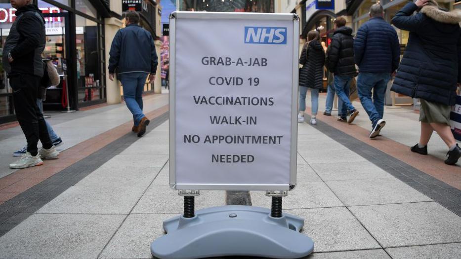 A sign in the middle of street that reads 'grab-a-jab Covid-19 vaccinations walk-in no appointment needed'. The sign has an NHS logo in the top right corner. Around the sign are lots of people walking past. 