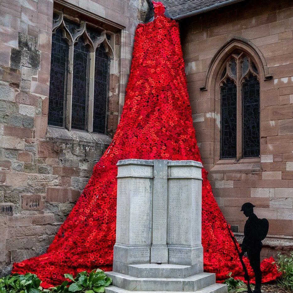 8,000 knitted poppies on the side of a church behind a grey memorial stone and a black shadow of a soldier holding a rifle.