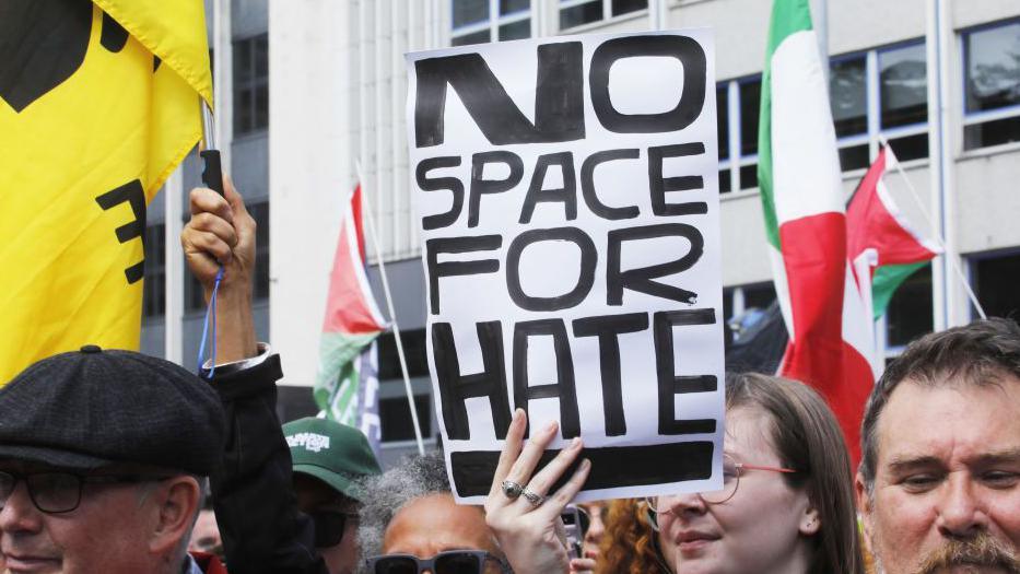 Anti-racism protestors. One is holding a sign saying "no space for hate"