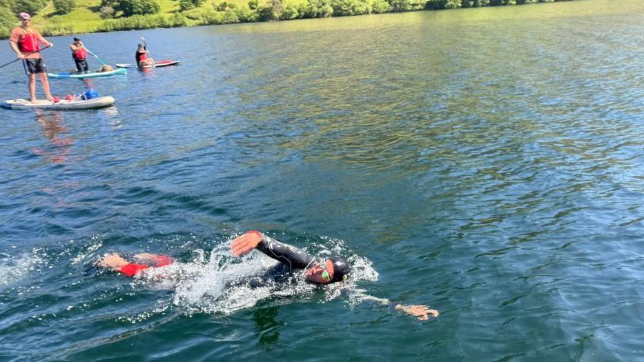 Adam Ball is swimming in Windermere. Three people are on paddle boards in the background watching.