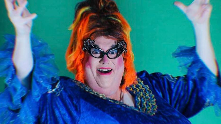 A pantomime dame, in the style of Dame Edna Everage, wearing a blue dress with bright orange hair and large glasses.