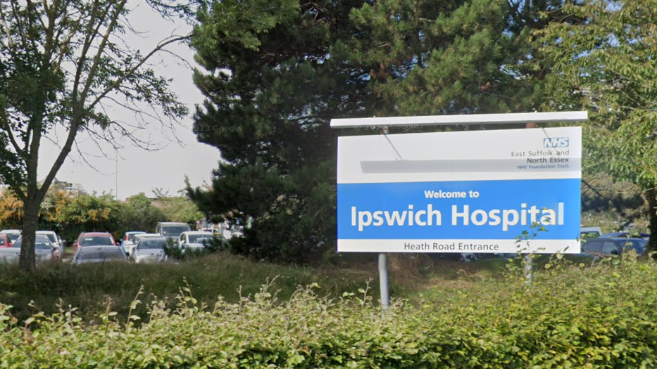 A blue-and-white sign saying "Welcome to Ipswich Hospital". There is a hedge in front of it and trees behind it. A car park can also be seen in the background.