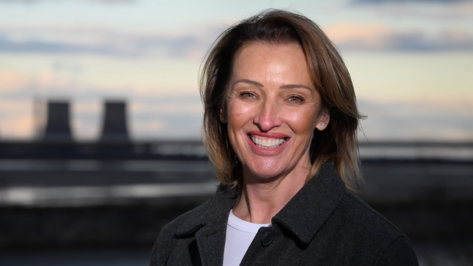 A publicity headshot of Sarah Pochin, smiling in a grey jacket with the power stations of Runcorn in the background