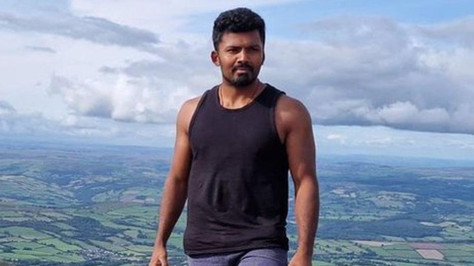 Mohananeethan Muruganantharajah is wearing a black vest-top and jeans while standing high on hillside overlooking a panoramic countryside view