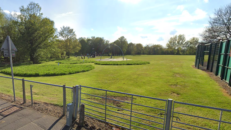 A view of Longwater Park in Costessey taken from the street. It shows a long area of grass and a children's play area in the background.