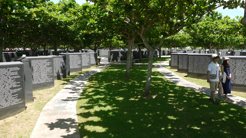 The photograph shows the Cornerstone of Peace Memorial in Japan which commemorates those lost during the Battle of Okinawa. Large granite blocks are arranged in rows in concentric rings and on each are dozens of names. A Japanese couple walk along the path gazing at the names. Many trees give shelter.