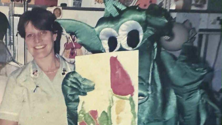 Trish Gwennap working at the children's ward at West Cornwall Hospital. She is stood to the left of a dressed up dragon character who is holding a painting. Trish has short dark hair and is smiling at the camera.