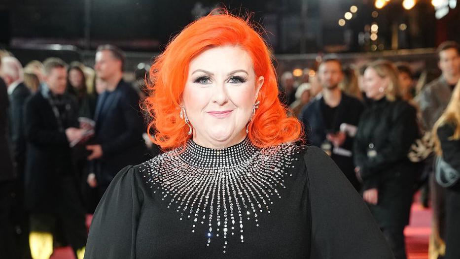 Michelle McManus smiles at the camera while on the red carpet. You can see lots of people in dark outfits blurred in the background behind her. The head and shoulders shot shows her orange, shoulder-length hair and black dress bedazzled with silver gemstones around her neck.