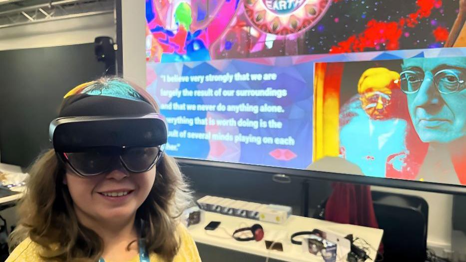 Beth Hands, a young female university student, wearing a VR headset as she stands in front of a colourful screen bearing an image of Gustav Holst