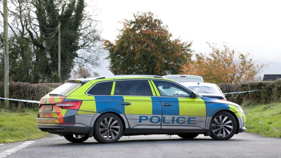 Yellow and blue police car at the scene of security alert in Keady, County Armagh. Car has a blue police logo on the side. There is police tape and trees and grass in the background.