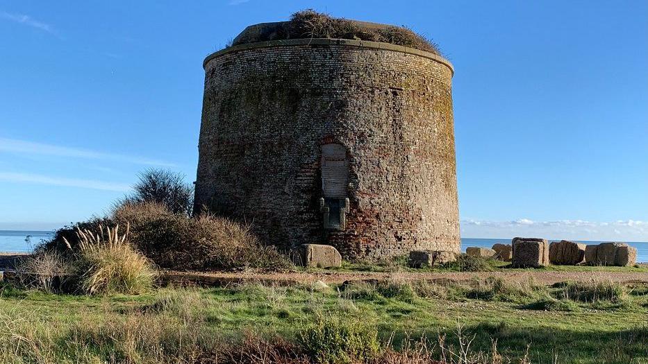 Martello Tower, a round brown stone building, surrounded by a few boulders and grasses, stands in front of and above the sea