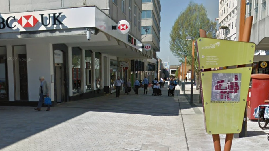 A green sign that reads "The Lexicon" with a map on. In the background there is a high street with shops.