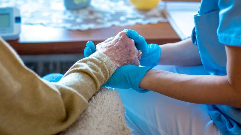 A female care worker wearing blue gloves hold the hand of an elderly lady sat in a chair