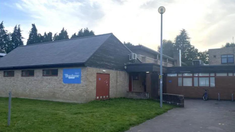 The front of Warminster Sports centre - a plain red brick building with a sign, a grassy area outside under a grey sky