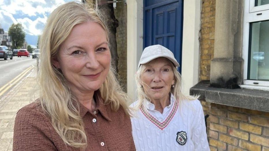 Laura Fraye and her mother Bonny Pitson are standing in front of a house. Laura has long blond hair and is wearing a brown blouse. Bonny also has long blond hair and is wearing a white cricket jumper and a white cap