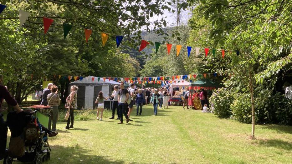 A grassy field with trees either side and crowds walking down the middle with a number of small marquees set up as stalls. There is colourful bunting hanging from the trees.