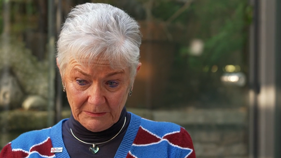 An elderly woman with wearing a blue jumper with red triangles on it. She is tearful and looking towards the ground