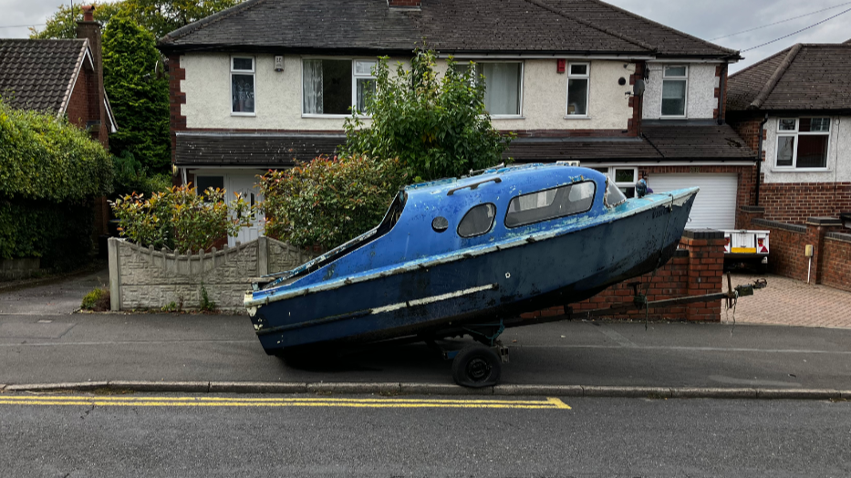 A side view of a blue boat on a trailer on the edge of a pavement, with semi-detached houses behind 