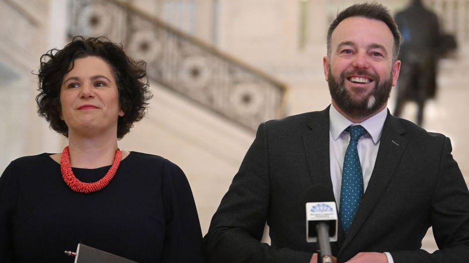 A man stands in front of a small black microphone as he buttons up his black suit. He has short brown hair and a short brown beard, with a small white patch on it. His shirt is white and his tie in blue with small white polka dots. To his right is a woman with short brown hair. She is wearing a black dress, thick red necklace. She has a smart watch on her left hand and a gold chain on her right, she holds a small black book. Behind the pair are white stairs with a black statue of a man at the top of them. 