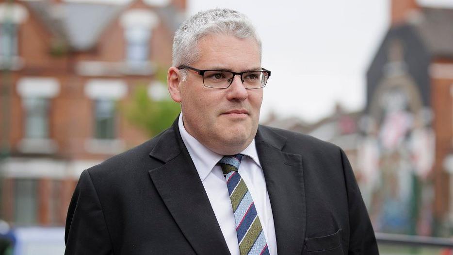 Gavin Robinson with short grey hair and black framed glasses wearing a black suit jacket, white shirt and striped tie.