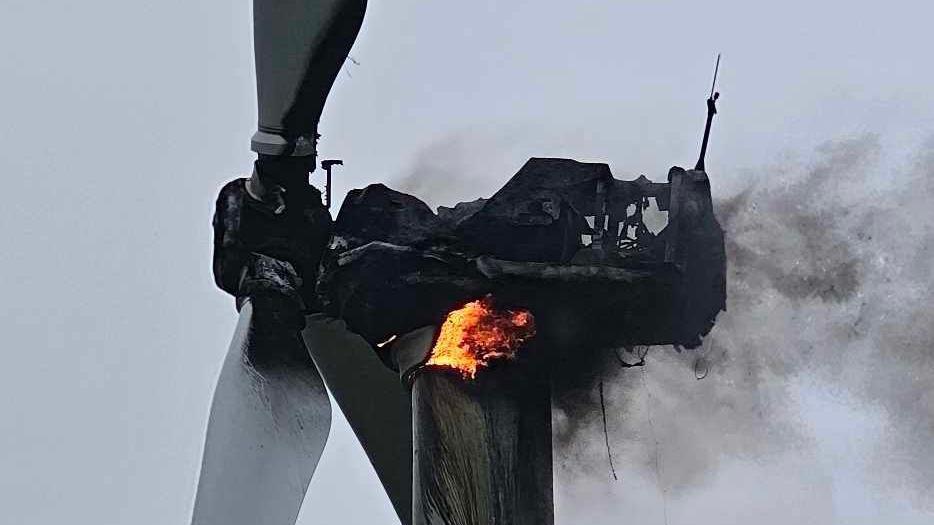 Close-up of the top of a wind turbine with red and orange flames at the centre. There is black smoke coming from the back of the wind turbine. The front has two white wings