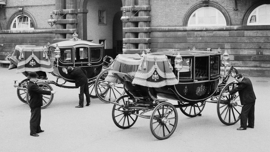 Two small coaches in front of an imposing stone building with workmen examining and cleaning the coaches while a bowler hatted supervisor looks on. 
