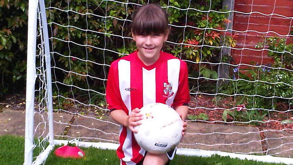 Olivia Clark, as a girl, smiling at the camera. She is kneeling and is holding a white football. She is wearing a red and white striped football shirt and is in front of a football goal.