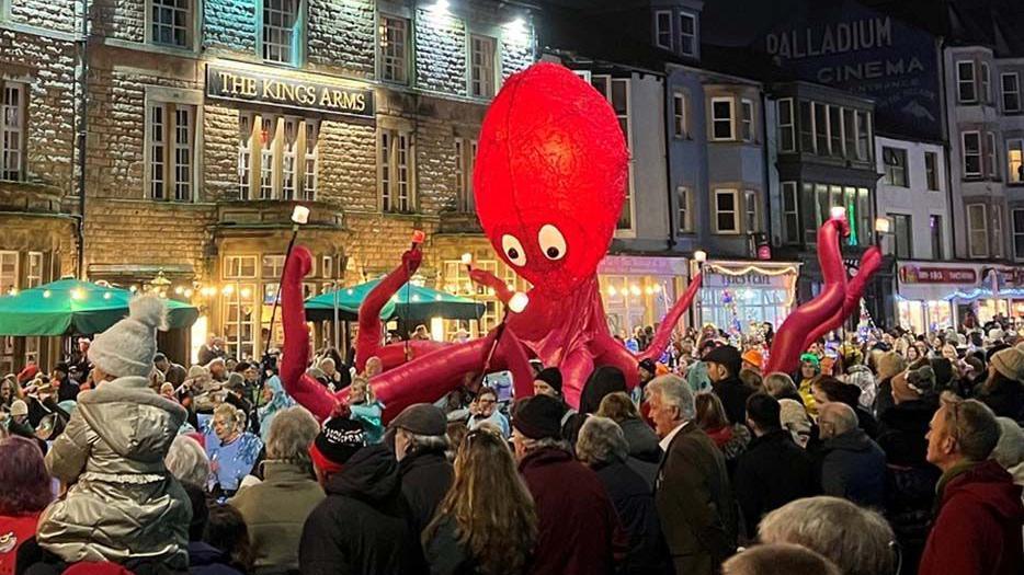 A red giant inflatable octopus with a glowing head being paraded through the streets of Morecambe surrounded by people.