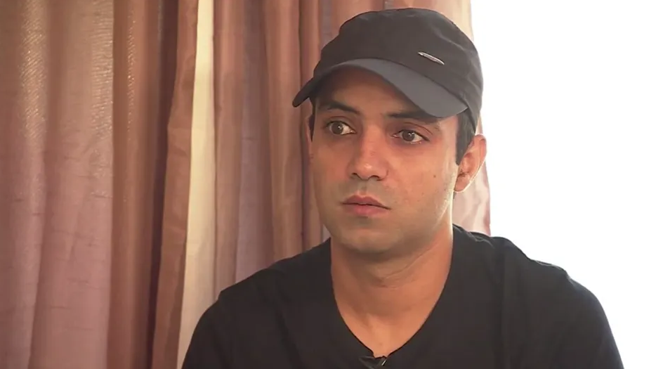 Nabeel Ahmad sat in front of a camera, wearing a black cap and black T-shirt, sat in front of a set of pink curtains 