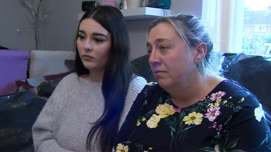 A young woman, possibly in her late teens with long, black hair, and a middle-aged woman with blonde hair up in a bun, sit side-by-side on a sofa, which is covered with a black plastic bin bag. The pair, who are mother and daughter, look concerned as they look off-camera.